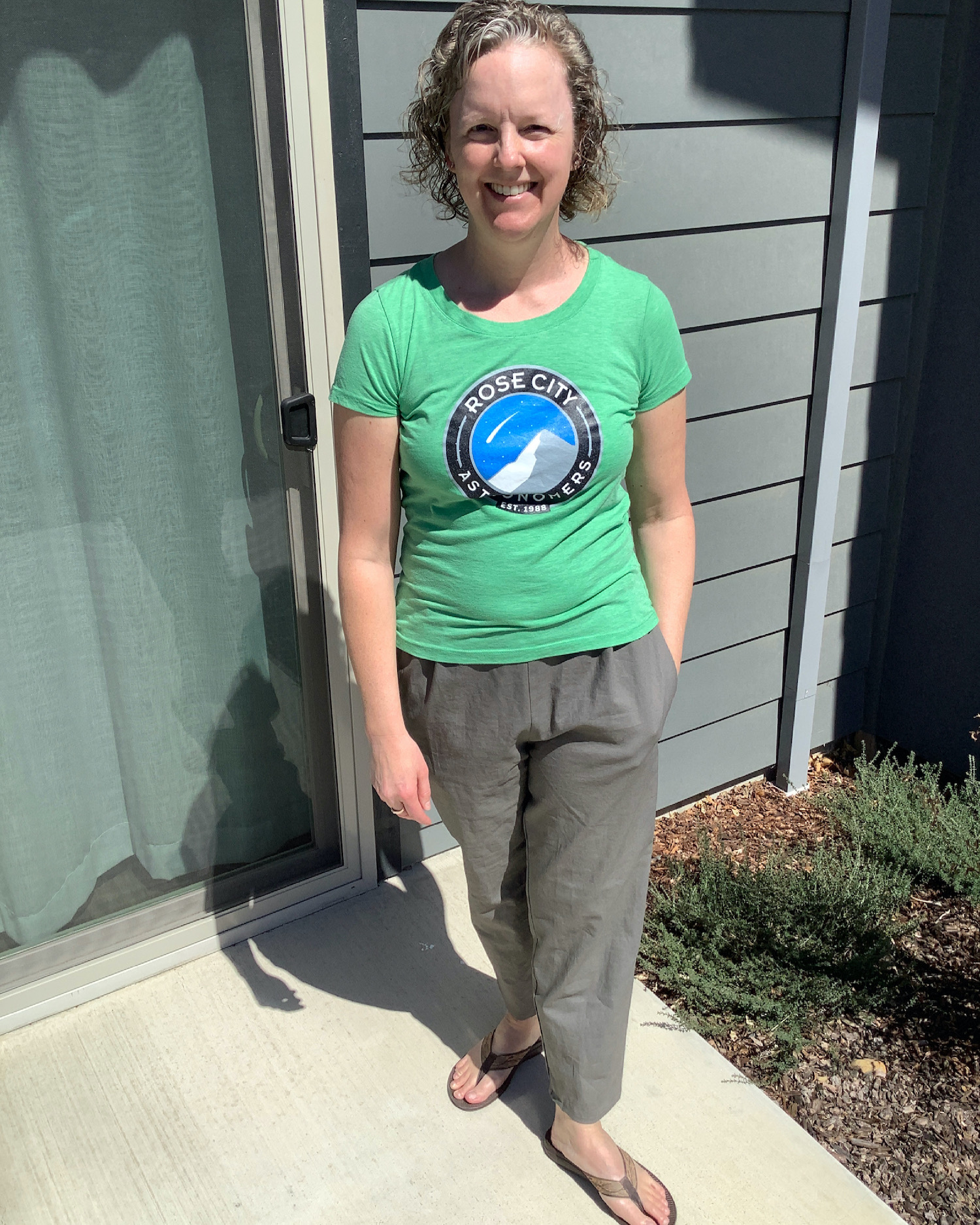 me standing in bright sunshine with a Rose City Astronomers t-shirt on and a pair of linen cropped pants, my eyes are squinting and I'm standing in front of a sliding glass door and the side of the house, next to a still hibernating plant with my left hand in my pocket.