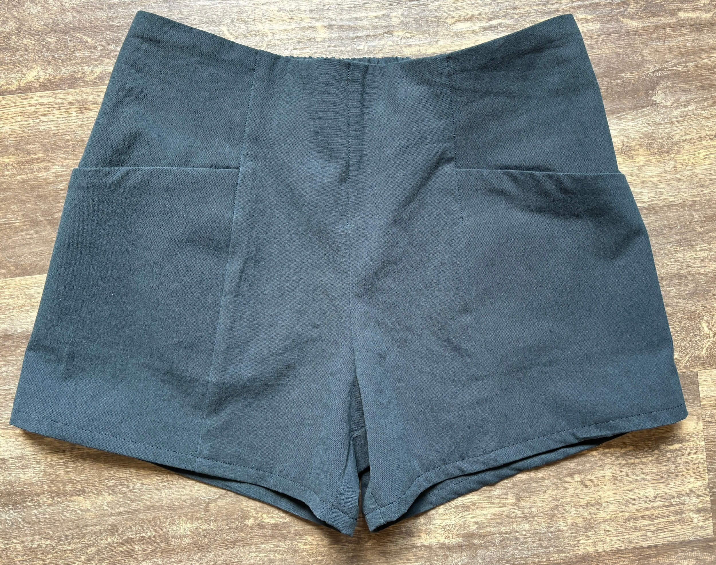 A pair of dark blue cotton shorts laying on a wood floor that is shades of brown. The shorts have an elastic band on the back waist and two pockets on the sides of the front that angle down.