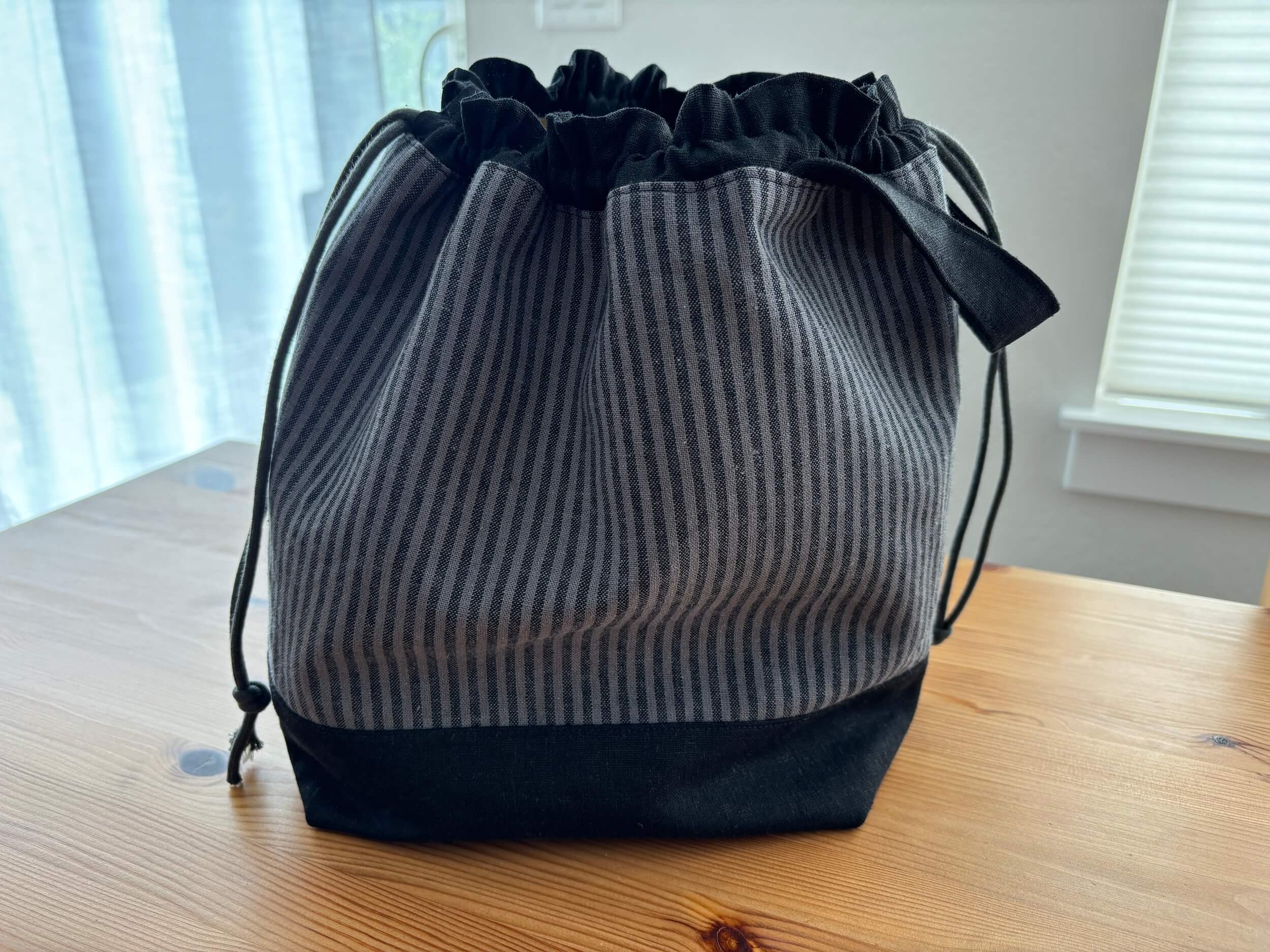 A bag with black on the top and a draw string, the middle section is a gray and black stripe and the bottom base is black. It's sitting on a table with a white wall behind it.