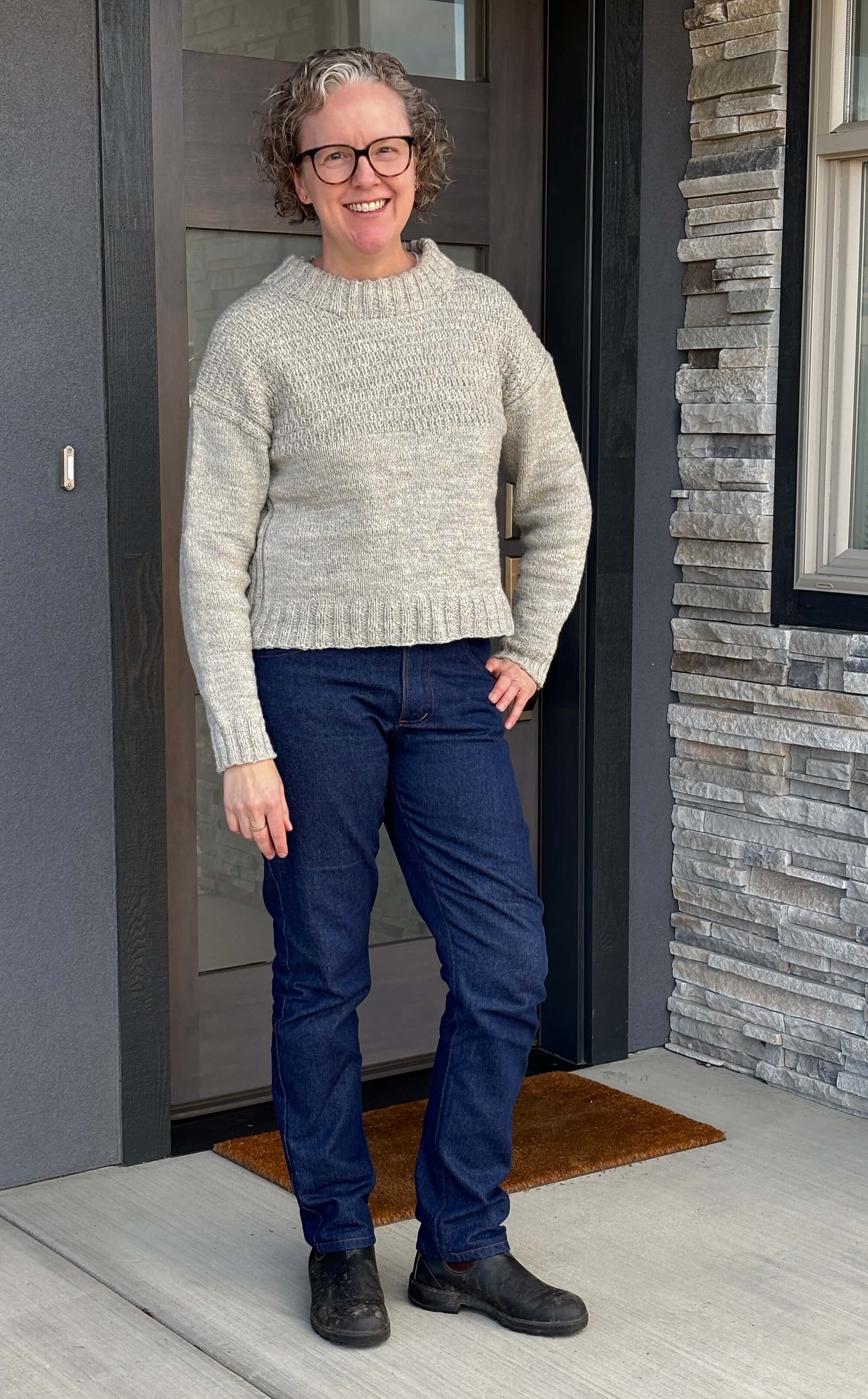 A woman stands in front of a doorway with a window to the left with a slight smile on her face. She has her left hand on her hop and is looking at the camera smiling. She's wearing a thick wool sweater with a texture on the top half in a neutral color and jeans in dark denim and dark boots. Her right arm rests at her side.