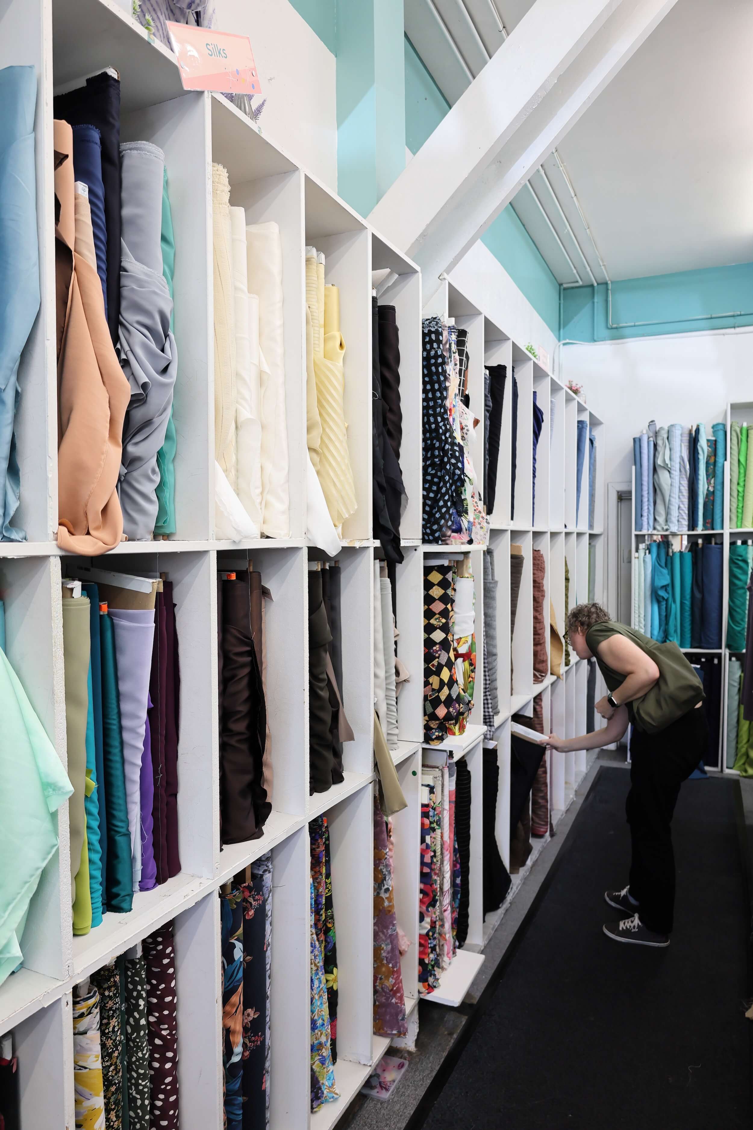 A wall of white shelving filled with bolts of fabric in all different colors and in the mid ground a woman stands bent down with a small bag over her shoulder pulling out a bolt of fabric to look more closely at it.