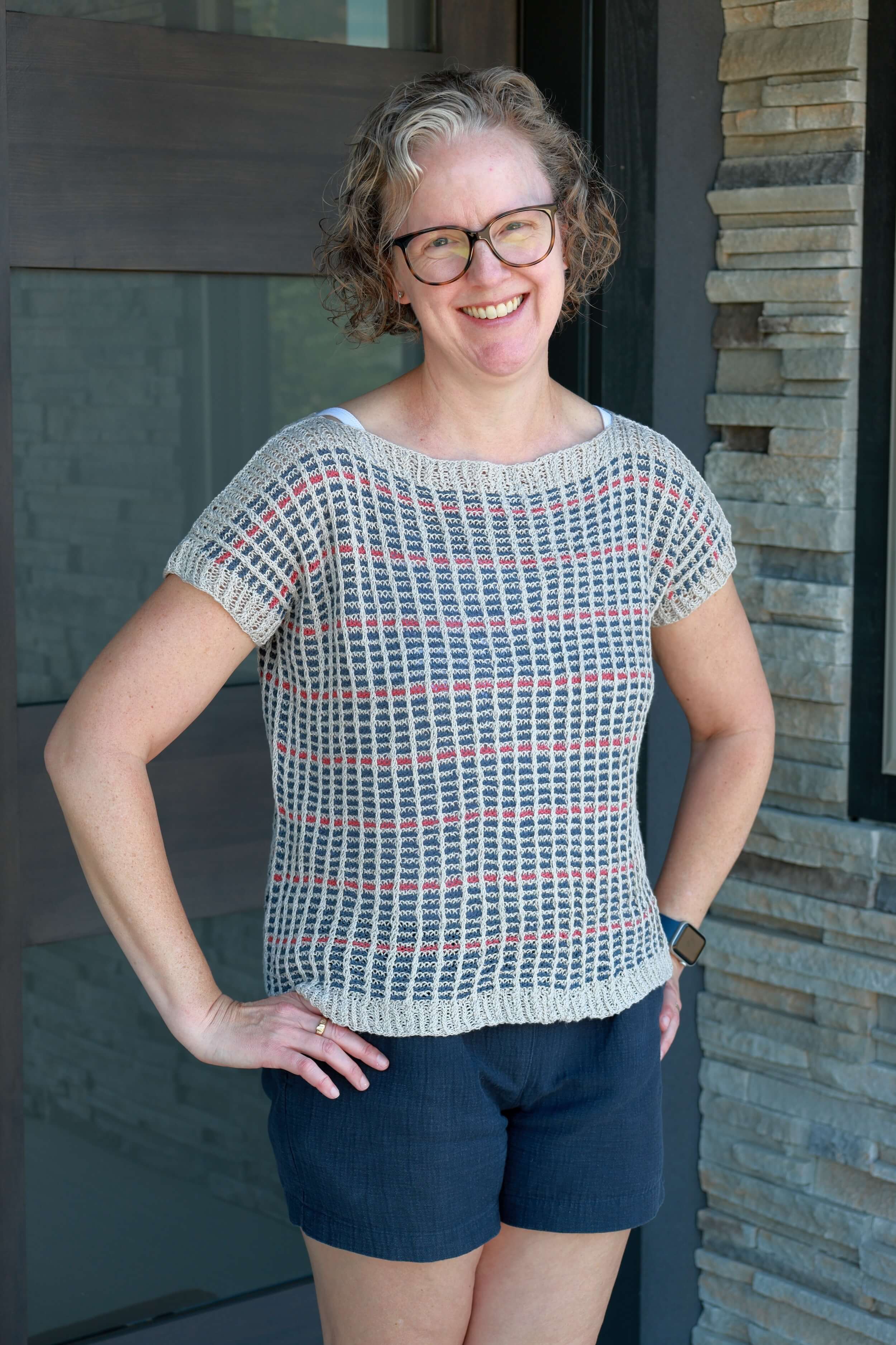 A woman stands with her hands on her hips in front of the front door of a house and a window that's next to it. She's got blond hair that's shoulder length and curly, wearing brown tortoise rim glasses, and is smiling. She's wearing a light gray boat neck knit top that has stripes of blue with a strip of a rasberry color every sixth stripe. She wears the knit with blue shorts.