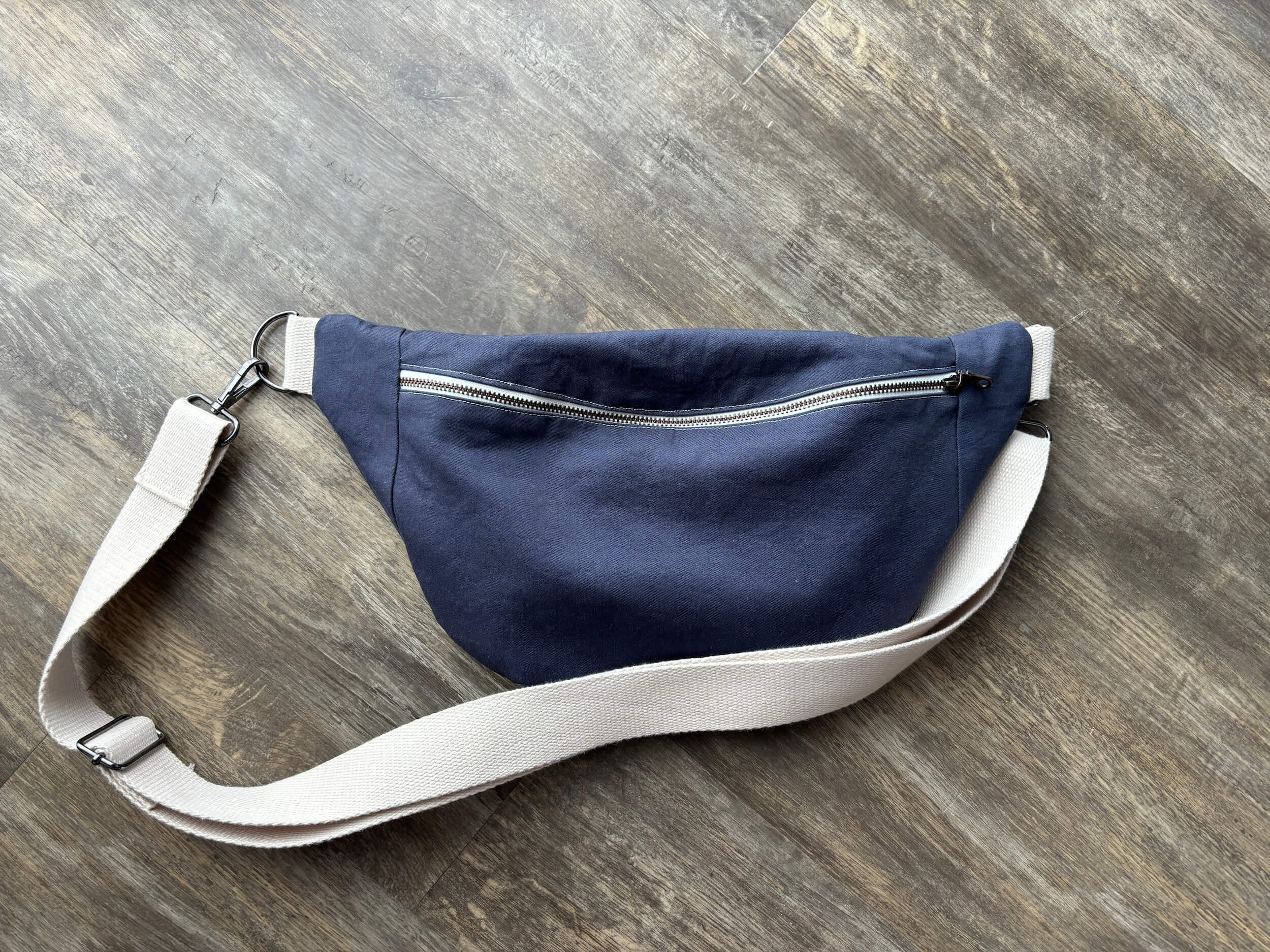 A blue bag with a white zipper laying on the floor with the cream colored strap laying over the top.