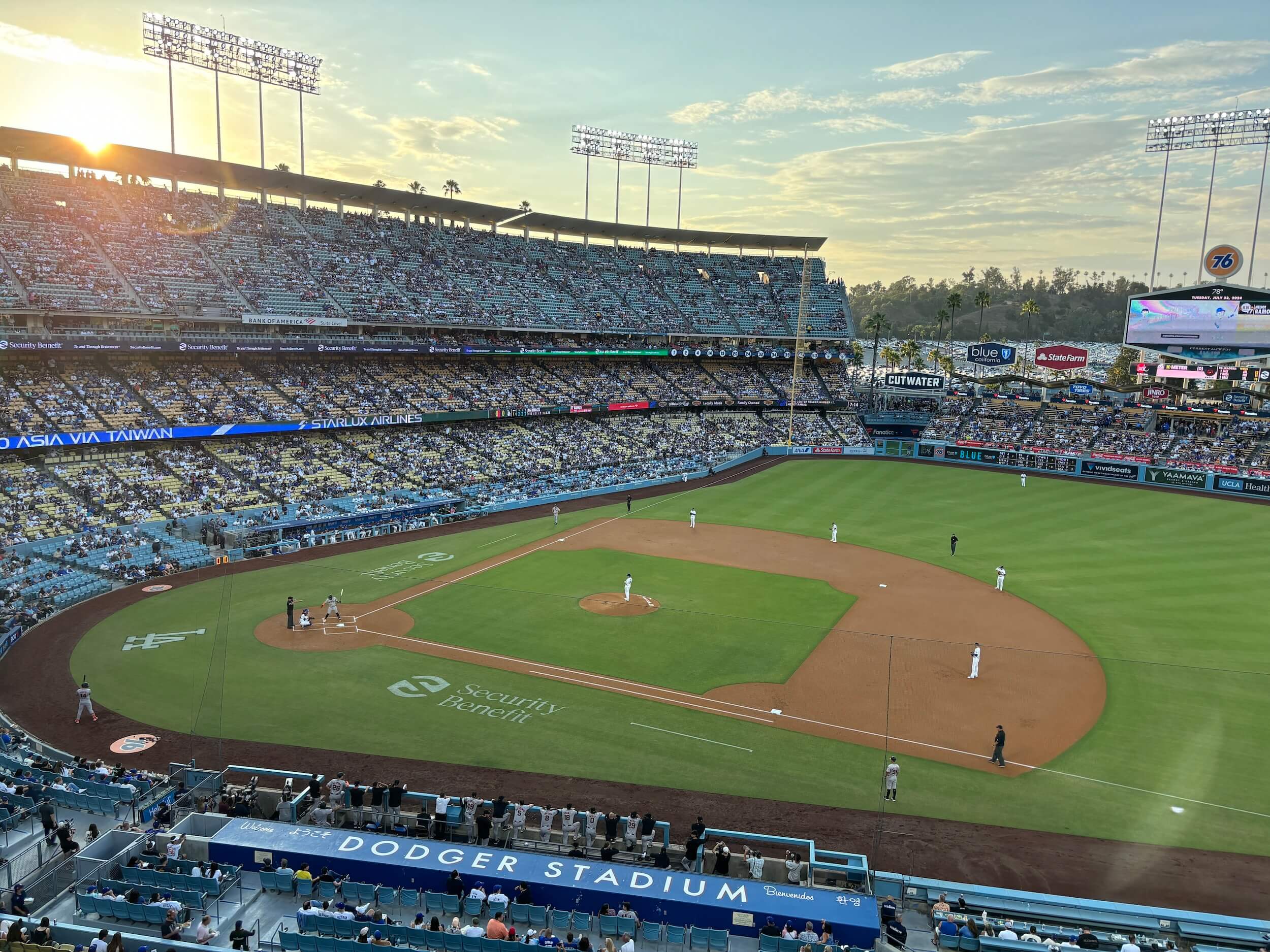 The sun is setting over the top of the stadium seating as baseball players on field play ball and the stands are full of people watching.