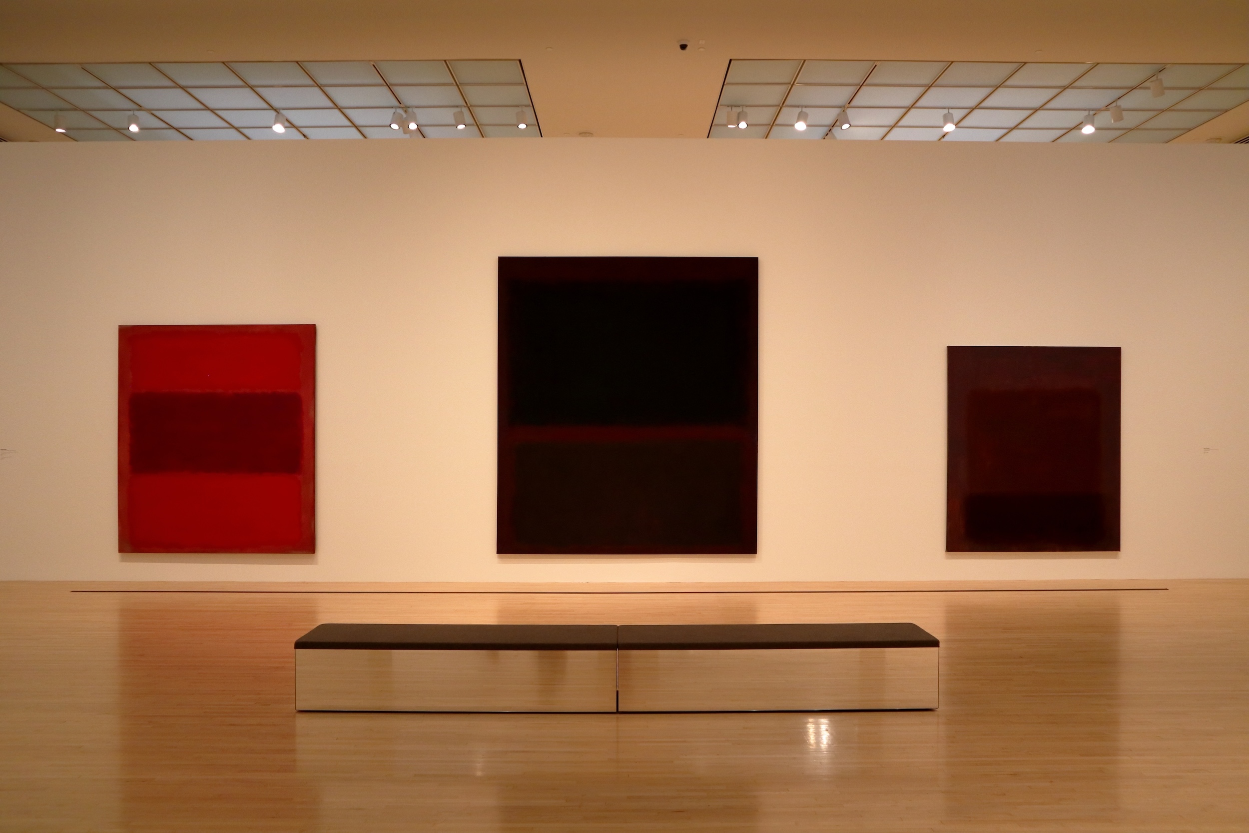 A wall of large paintings that are color fields, there are three with the one on the left being rectangeles of red, the one in the middle deep purple almost black in areas, and the one on the right deep to lighter purple shades. There is an empty bench in front of the paintings.